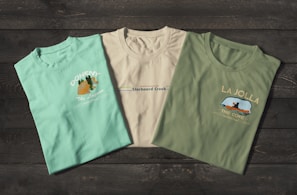 Three t-shirts are displayed on a dark wooden surface. The t-shirts have different prints, with the first one being light turquoise with the word 'Doheny', an orange fish graphic, and the text 'The Lagoon'. The second shirt is off-white featuring 'Starboard Creek' across the chest. The third t-shirt is olive green with the words 'La Jolla' and an image of a kayak with 'The Cove' written underneath.