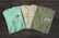 Three t-shirts are displayed on a dark wooden surface. The t-shirts have different prints, with the first one being light turquoise with the word 'Doheny', an orange fish graphic, and the text 'The Lagoon'. The second shirt is off-white featuring 'Starboard Creek' across the chest. The third t-shirt is olive green with the words 'La Jolla' and an image of a kayak with 'The Cove' written underneath.