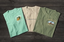 Three t-shirts are displayed on a dark wooden surface. The t-shirts have different prints, with the first one being light turquoise with the word 'Doheny', an orange fish graphic, and the text 'The Lagoon'. The second shirt is off-white featuring 'Starboard Creek' across the chest. The third t-shirt is olive green with the words 'La Jolla' and an image of a kayak with 'The Cove' written underneath.