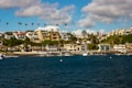 A coastal area features a collection of luxury homes set on a hillside. The houses vary in architectural style and are surrounded by lush greenery and tall palm trees. Below the homes, a marina is visible with several boats docked along the wooden piers extending into deep blue water. The sky above is partly cloudy, creating a relaxed and inviting atmosphere.