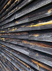 A close-up view of weathered wooden planks arranged horizontally. The wood has darkened with age and exposure, with hints of golden and amber tones highlighting the grain patterns and knots.