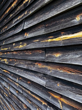A close-up view of weathered wooden planks arranged horizontally. The wood has darkened with age and exposure, with hints of golden and amber tones highlighting the grain patterns and knots.