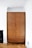 A wooden wardrobe stands against a white wall in a room with parquet flooring. It features a simple, minimalistic design with a medium brown finish. To the side, there is part of a black fireplace visible. The room's corner ceiling details are visible, highlighting a clean, modern aesthetic.