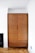 A wooden wardrobe stands against a white wall in a room with parquet flooring. It features a simple, minimalistic design with a medium brown finish. To the side, there is part of a black fireplace visible. The room's corner ceiling details are visible, highlighting a clean, modern aesthetic.