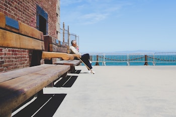 A person is sitting alone on a wooden bench alongside a brick wall, gazing towards a fenced waterfront with a clear blue sky. The scene conveys a sense of solitude and contemplation.