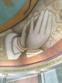 A large mural depicting a pair of hands in a prayer gesture, adorned with beads, set against a decorative background. The mural is situated on a curved ceiling or wall surface, accompanied by a Latin inscription. The architectural elements include columns and a detailed trim.