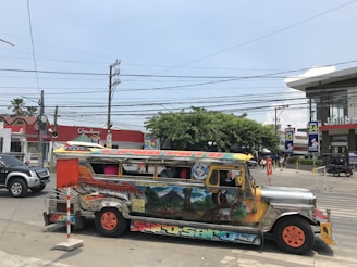 A brightly painted jeepney with various colors and illustrations is parked on a busy street. The jeepney features designs and text, including 'Saro-Saro'. The background includes a Chowking and a Jollibee, both fast-food restaurants, with visible signage. There are several vehicles on the road, including a black SUV. There are also numerous power lines overhead and some trees providing greenery.