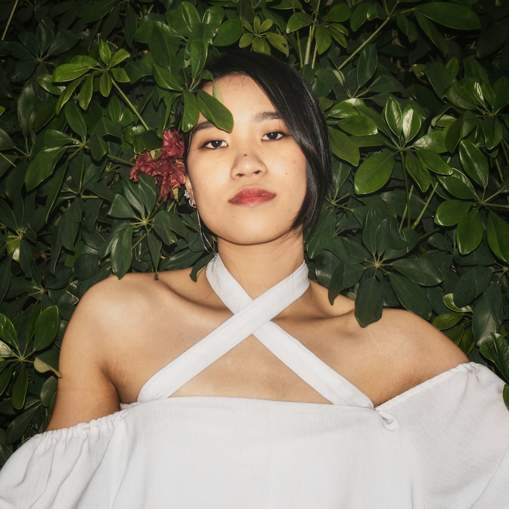 woman in white top with flower on right ear leaning to a green plant