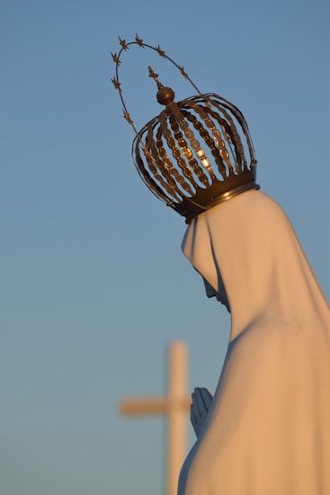 A statue of a praying figure wearing a decorative crown, set against a clear blue sky with a blurred cross in the background.