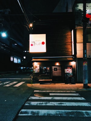 A small, dimly lit restaurant with a wooden exterior is visible at a street corner. The illuminated signage displays Japanese characters, and the entrance is decorated with posters and a menu board. A bicycle is partially visible to the side, and a zebra crossing is in the foreground.