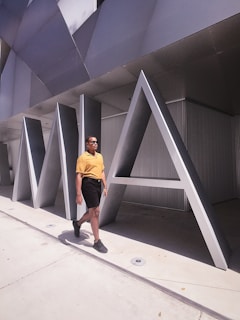 A person wearing a yellow shirt and black shorts walks confidently beside large metallic architectural letters forming the word 'MIA'. The setting appears to be outdoors in a modern, urban environment.
