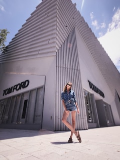 A modern, geometric building with a textured facade stands against a clear sky. A fashionable person wearing a denim outfit and sunglasses leans casually in front of the building, creating a stylish and urban atmosphere.