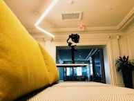 A modern open space featuring a yellow couch in the foreground, a camera mounted on a gimbal positioned centrally on a table, and an exit sign on a white ornate ceiling. The room is well-lit with LED lighting and there is a large mirror or glass wall reflecting part of the interior. A plant is visible to the side.