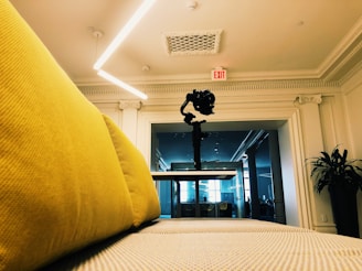 A modern open space featuring a yellow couch in the foreground, a camera mounted on a gimbal positioned centrally on a table, and an exit sign on a white ornate ceiling. The room is well-lit with LED lighting and there is a large mirror or glass wall reflecting part of the interior. A plant is visible to the side.