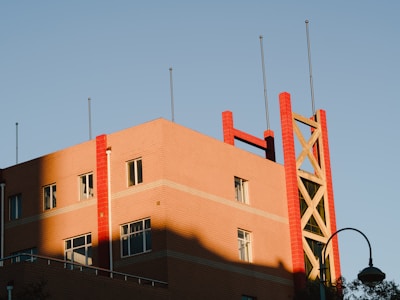 A building with a modern architectural design features red brick accents and geometric patterns. Tall metal poles protrude from the roof, and the structure is partially illuminated by sunlight, creating shadows across the facade. The sky is clear and blue, providing a crisp backdrop.