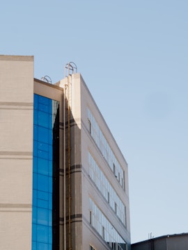 A modern building with a facade of large glass windows and beige brickwork. The structure has metal ladders attached to the side leading to the roof, and the clear blue sky provides a contrasting backdrop.