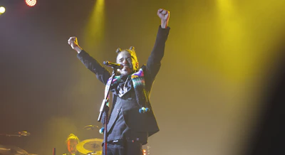 A performer on stage holds a microphone with both arms raised triumphantly. The lighting is dramatic with yellow spotlighting and a dark background. The performer wears a dark suit accented with colorful elements.