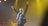 A performer on stage holds a microphone with both arms raised triumphantly. The lighting is dramatic with yellow spotlighting and a dark background. The performer wears a dark suit accented with colorful elements.