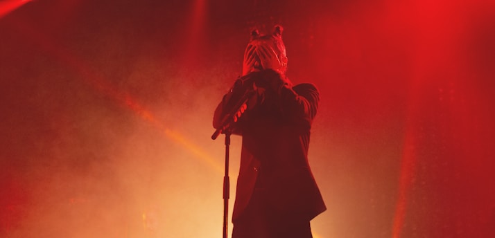 A person stands on stage, holding a microphone stand, wearing a mask and appearing to be performing or delivering a speech. The scene is saturated with red and orange lighting, creating a dramatic and intense atmosphere.