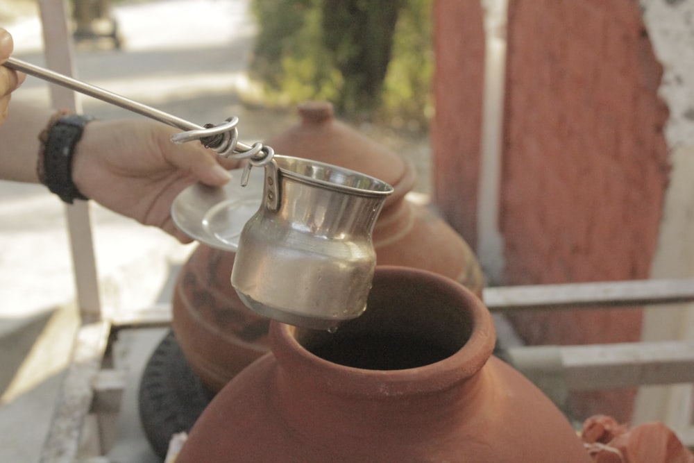 person holding gray stainless steel mini jar with handle near brown stone jar