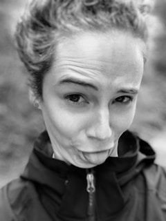 A person making a playful facial expression, sticking out their tongue and raising their eyebrows. The image is in black and white, and the person is wearing a dark jacket.
