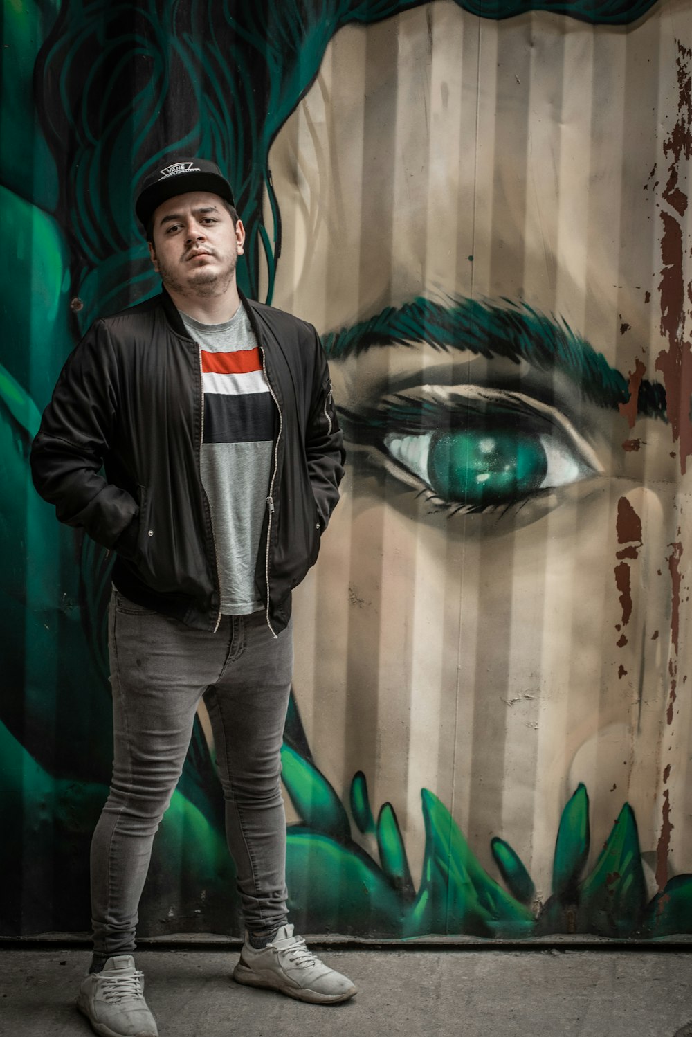 man with both hands in his jacket's pocket standing in front of wall with woman mural