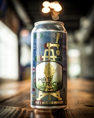 A can of beer is prominently displayed on a wooden surface, featuring a label with a gold illustration of a tower and a hop. Various colorful symbols resembling flags are scattered across the design. The background is blurred, with soft lighting creating a warm atmosphere.