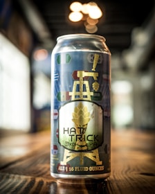 A can of beer is prominently displayed on a wooden surface, featuring a label with a gold illustration of a tower and a hop. Various colorful symbols resembling flags are scattered across the design. The background is blurred, with soft lighting creating a warm atmosphere.