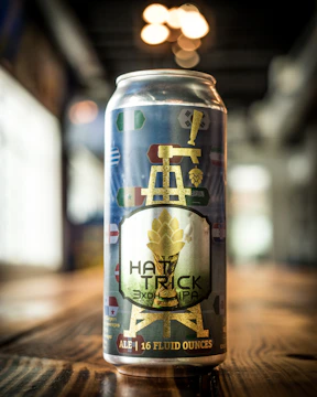 A can of beer is prominently displayed on a wooden surface, featuring a label with a gold illustration of a tower and a hop. Various colorful symbols resembling flags are scattered across the design. The background is blurred, with soft lighting creating a warm atmosphere.