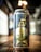A can of beer is prominently displayed on a wooden surface, featuring a label with a gold illustration of a tower and a hop. Various colorful symbols resembling flags are scattered across the design. The background is blurred, with soft lighting creating a warm atmosphere.