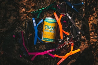 A variety of colorful sunglasses arranged in a circular pattern around a can of beer labeled 'Cheap Sunglasses' from Bakfish Brewing Co. The arrangement is set on a dark, natural surface that appears to be a tree stump, surrounded by dried leaves.