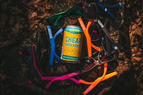 A variety of colorful sunglasses arranged in a circular pattern around a can of beer labeled 'Cheap Sunglasses' from Bakfish Brewing Co. The arrangement is set on a dark, natural surface that appears to be a tree stump, surrounded by dried leaves.