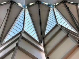 Angular architectural structure with large triangular glass panels separated by metal beams and hinges. Natural light enters through the glass, highlighting the geometric design and industrial style.