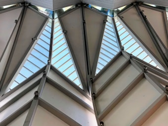 Angular architectural structure with large triangular glass panels separated by metal beams and hinges. Natural light enters through the glass, highlighting the geometric design and industrial style.