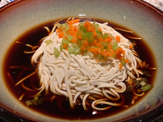 A bowl filled with dark broth containing a generous serving of pale, freshly cooked noodles. The noodles are topped with finely chopped green and orange vegetables.