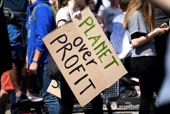 Several people are gathered, with one prominently holding a cardboard sign reading 'Planet over Profit'. They appear to be at a rally or protest, emphasizing a message focused on environmental concerns. The scene is bright with sunlight, and the participants are casually dressed.