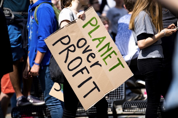 Several people are gathered, with one prominently holding a cardboard sign reading 'Planet over Profit'. They appear to be at a rally or protest, emphasizing a message focused on environmental concerns. The scene is bright with sunlight, and the participants are casually dressed.