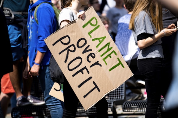 Several people are gathered, with one prominently holding a cardboard sign reading 'Planet over Profit'. They appear to be at a rally or protest, emphasizing a message focused on environmental concerns. The scene is bright with sunlight, and the participants are casually dressed.