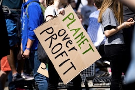 Several people are gathered, with one prominently holding a cardboard sign reading 'Planet over Profit'. They appear to be at a rally or protest, emphasizing a message focused on environmental concerns. The scene is bright with sunlight, and the participants are casually dressed.