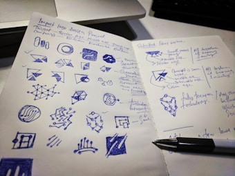 A two-page notebook spread filled with hand-drawn sketches and notes. The left side contains various geometric shapes and logo designs drawn in blue ink, while the right page has handwritten notes and selected ideas with a pen resting on it. A computer keyboard is partially visible in the background.