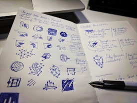 A two-page notebook spread filled with hand-drawn sketches and notes. The left side contains various geometric shapes and logo designs drawn in blue ink, while the right page has handwritten notes and selected ideas with a pen resting on it. A computer keyboard is partially visible in the background.