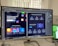 A computer monitor displays a graphic design software interface, specifically Adobe Photoshop CC 2018. The screen shows a digital webpage layout with various sections, featuring vibrant and colorful graphics. In the foreground, a desk with a notebook and some handwritten notes is visible, while another partially visible screen displays a scenic green landscape.