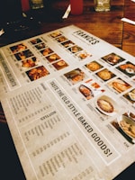 A restaurant menu placed on a wooden table, featuring images and prices of various dishes and drinks. The menu includes sections for starters, main courses, and beverages, with detailed photographs of each item. Items like baked goods, pasta, and drinks are highlighted.