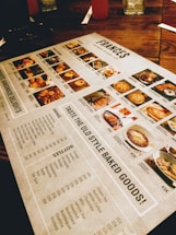 A restaurant menu placed on a wooden table, featuring images and prices of various dishes and drinks. The menu includes sections for starters, main courses, and beverages, with detailed photographs of each item. Items like baked goods, pasta, and drinks are highlighted.
