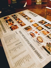 A restaurant menu placed on a wooden table, featuring images and prices of various dishes and drinks. The menu includes sections for starters, main courses, and beverages, with detailed photographs of each item. Items like baked goods, pasta, and drinks are highlighted.