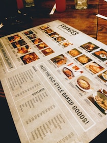 A restaurant menu placed on a wooden table, featuring images and prices of various dishes and drinks. The menu includes sections for starters, main courses, and beverages, with detailed photographs of each item. Items like baked goods, pasta, and drinks are highlighted.