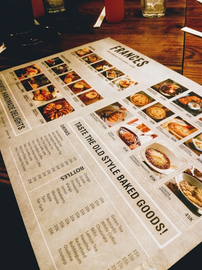 A restaurant menu placed on a wooden table, featuring images and prices of various dishes and drinks. The menu includes sections for starters, main courses, and beverages, with detailed photographs of each item. Items like baked goods, pasta, and drinks are highlighted.