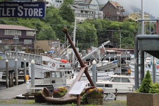 A harbor scene with several boats docked at a marina surrounded by lush green hills and various buildings. A large anchor is placed prominently in the foreground with a small flowering planter beside it. The setting appears to be a quaint coastal town with a mix of residential and commercial buildings.