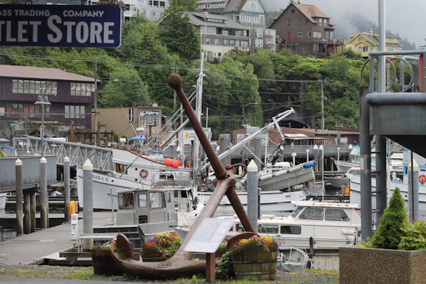 A harbor scene with several boats docked at a marina surrounded by lush green hills and various buildings. A large anchor is placed prominently in the foreground with a small flowering planter beside it. The setting appears to be a quaint coastal town with a mix of residential and commercial buildings.