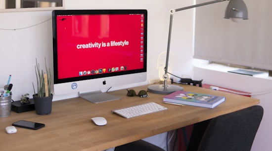 A modern workspace featuring a desktop computer with a bold red wallpaper displaying the phrase 'creativity is a lifestyle'. The desk is organized with a potted plant, books, a keyboard, and a mouse. Nearby, a sleek desk lamp provides additional lighting, and personal items like glasses and a smartphone are neatly placed.